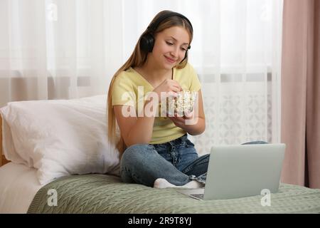 Teenage girl with headphones eating popcorn while using laptop on bed at home Stock Photo