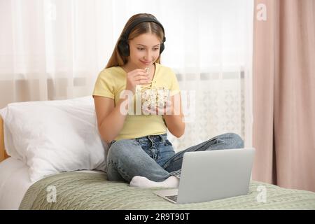 Teenage girl with headphones eating popcorn while using laptop on bed at home Stock Photo