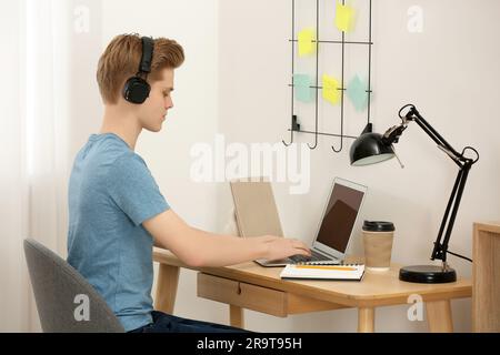 Teenage boy with headphones using laptop at wooden desk in room Stock Photo
