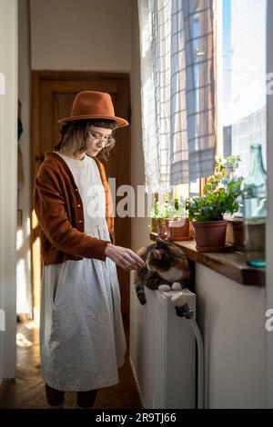 Artist girl in hat stands near window enjoys rays sun, plays with fluffy cat lying on radiator. Stock Photo