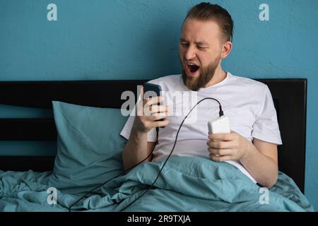Handsome man connects his smartphone to a power bank while lying on the bed at home, waking up in the morning, charging the gadget Stock Photo