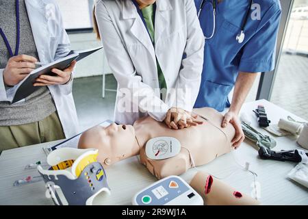 first aid seminar, partial view of young woman in white coat practicing chest compressions on CPR manikin near paramedic and student writing in notebo Stock Photo