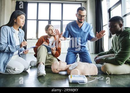 professional paramedic gesturing and talking to multiethnic participants near CPR manikin and defibrillator during first aid seminar in training room, Stock Photo