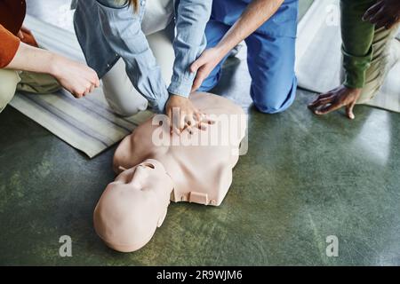 cropped view of paramedic assisting young woman practicing chest compressions on CPR manikin near multiethnic participants of first aid training semin Stock Photo