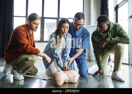 young asian woman doing chest compressions on CPR manikin while practicing cardiopulmonary resuscitation near paramedic and multiethnic participants, Stock Photo