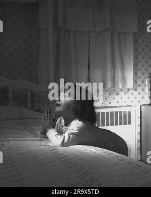 Little girl kneeling next to her bed in her pajamas, leaning on the bed with her hands together praying while looking up Stock Photo