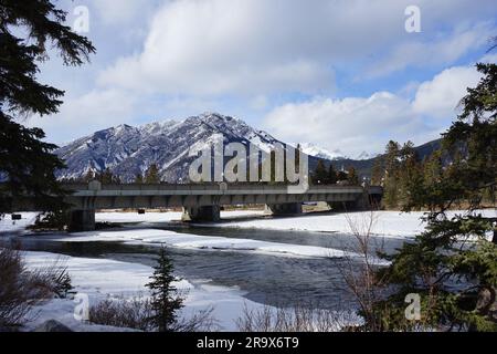 A scenic winter landscape featuring a bridge crossing over a snow-covered river surrounded by mountains in the background Stock Photo