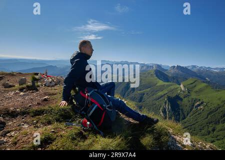 man with backpack on top of a hill with mountains, landscape. man sitting on grass overlooking mountains Stock Photo