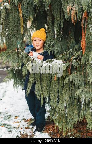Happy little boy playint with snow outside, wearing warm winter jacket and yellow hat. Child playing snowballs, hiding behind the tree Stock Photo