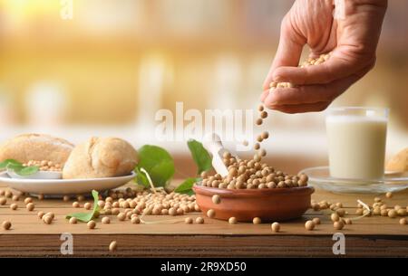 Hand dropping soy beans on wooden table with soy products and kitchen in the background. Front view. Stock Photo