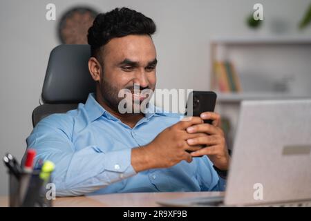 Happy smiling indian corporate employee using mobile phone in front of laptop at office - concept of break time, relaxation and technology. Stock Photo