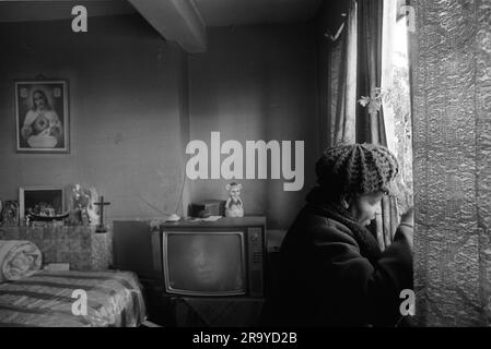 East End London 1970s UK poverty in Britain, an elderly black British woman in her council estate flat on the Stonebridge Estate.  Isolated and living alone in a single room plus small kitchen and bathroom. All in very bad condition, the flat is damp and very cold, she is dressed in her hat and coat to keep warm. On her bed is a 'bedroll' above her bed an image of Christ. Haggerston, East London, England 1978. 70s HOMER SYKES Stock Photo