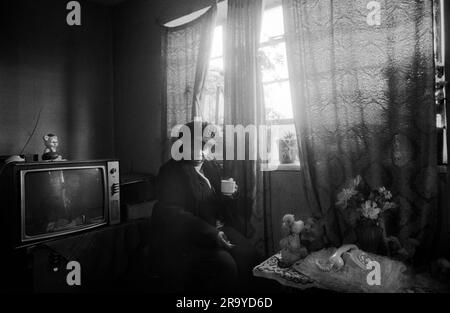 East End London 1970s, poverty in Britain, an elderly black British woman in her council estate flat on the Stonebridge Estate.  Isolated and living alone in a single room plus small kitchen and bathroom. All in very bad condition, the flat is damp and very cold, she is dressed in her hat and coat and drinks a cup of tea to keep warm. Haggerston, East London, England 1978. 70s UK HOMER SYKES Stock Photo