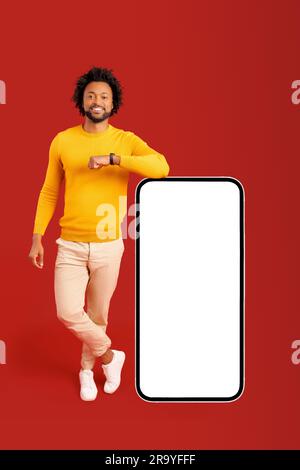 Cheerful confident African-American young man is leaning at huge smartphone with empty screen isolated on red background, curly guy with toothy smile presenting and advertising mobile app, full length Stock Photo