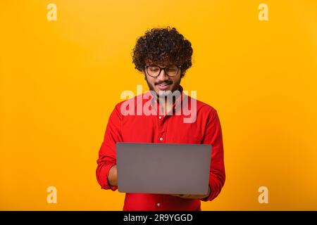 Portrait of cheerful young brunette man with beard typing email on laptop and smiling, reading positive message or surfing the web. Indoor studio shot isolated on orange background Stock Photo