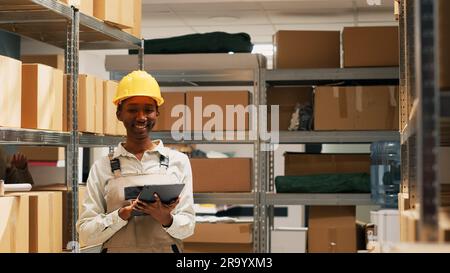African american woman holding tablet to do inventory in storage room space, using device to check merchandise stock before shipping products. Female worker planning order distribution. Stock Photo