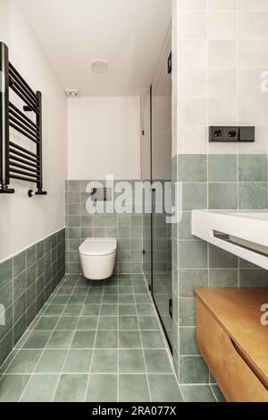 A newly installed modern bathroom with a hanging toilet, white porcelain sink, walk-in shower and a black wall-mounted towel rail Stock Photo