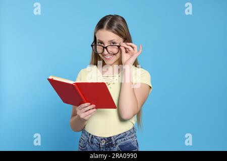 Teenage girl in eyeglasses reading book on light blue background Stock Photo