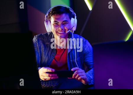 Gamer smiling at the camera with a headset on and a smartphone in his hand. Sitting at his gaming station, a young male player enjoys watching a live Stock Photo