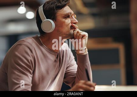 Designer thinking about his project while sitting in a cafe. Male graphic design professional wearing wireless headphones. Stock Photo