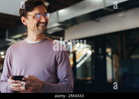 Pensive business man smiling using a cellphone in a modern workplace. Happy business man texting on a mobile phone while standing in an office. Stock Photo