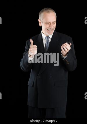 An amiable businessman wearing a black suit smiling and reading on mobile phone, with thumb up, on black background Stock Photo