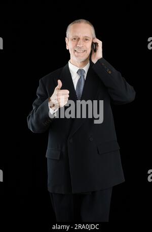 An amiable businessman wearing a black suit smiling and speaking on mobile phone, with thumb up, on black background Stock Photo