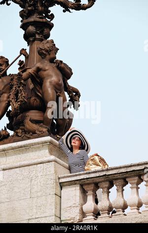 A glamourous woman in Paris on Pont Alexandre III bridge leans against the balustrade under the bronze cupid sculpture of the ornate Art Nouveau lamps Stock Photo