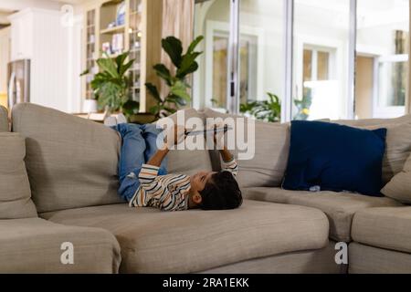 Happy biracial boy lying on couch with feet up using tablet in living room, copy space Stock Photo