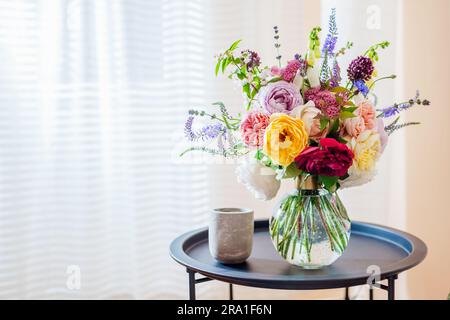 Close up of summer bouquet of roses flowers put in transparent vase on table by window. Cozy interior and decor at home. Space Stock Photo