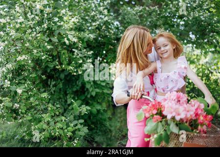 Young attractive mother rides a bicycle with her cute little daughter. A charming blonde woman and a cheerful laughing girl are having fun. A flowerin Stock Photo