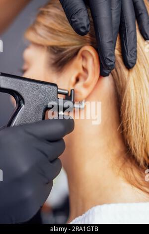 Close up of face of young blonde woman doing ear piercing procedure. Doctor hands in black gloves hold ear piercing gun. Medical worker making hole an Stock Photo