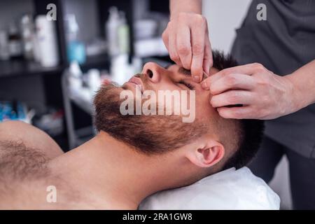 Close up portrait of a young attractive man getting facial massage. Masseuse hands on the male client chin. Brunette girl closed her eyes and enjoys a Stock Photo