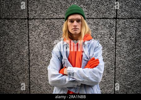 European millennial hipster guy looking at camera, standing with crossed arms against gray wall  Stock Photo