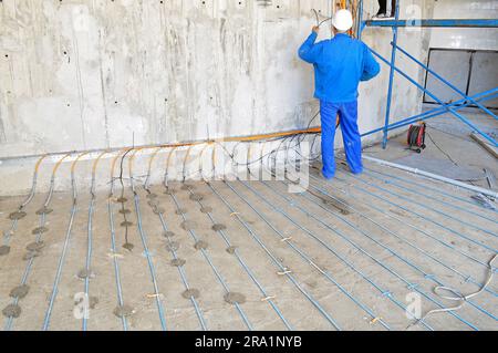 A man wearing a blue uniform is seen installing an underfloor heating cable on a concrete surface, captured from behind Stock Photo