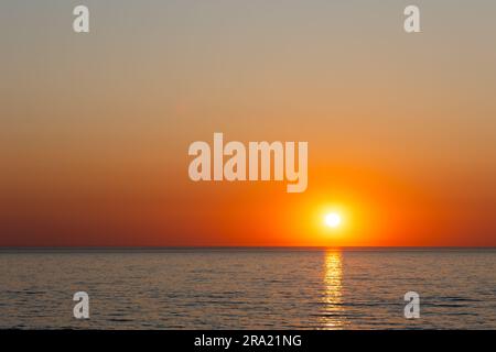 A large orange sun setting over a calm sea. Static shot taken during a cloudless evening Stock Photo