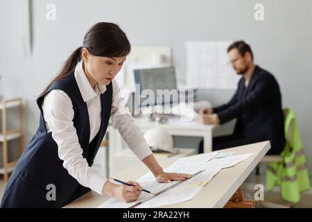 Side view portrait of young woman as engineer drawing blueprints and plans at table in office, copy space Stock Photo