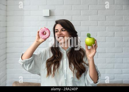 Young student woman doubts what to choose healthy food or sweets junk unhealthy food holding green apple and donut in hands standing at home in her li Stock Photo