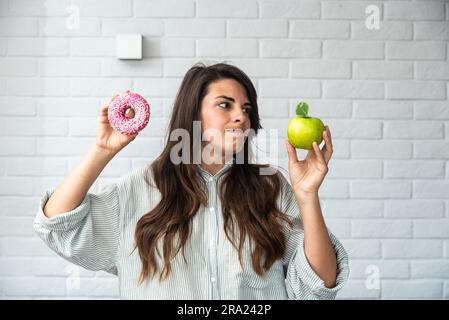 Young student woman doubts what to choose healthy food or sweets junk unhealthy food holding green apple and donut in hands standing at home in her li Stock Photo