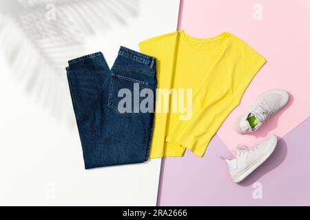 Yellow t-shirt, blue jeans and white sneakers on pink and white background with palm leaf shadow. Directl above, high angle shot. Stock Photo