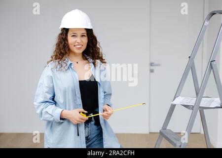 repair, construction and building concept - woman in goggles and protective gloves with ruler and pencil measuring wall at home. Stock Photo
