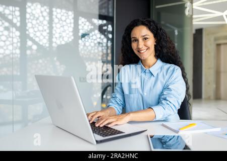 Young beautiful Indian female programmer working inside the office on a laptop, woman smiling and looking at the camera, portrait of a satisfied and confident businesswoman at the workplace. Stock Photo