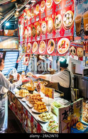 Woman serving young woman at a Chinese food stall selling foods on a stick, dumplings and steamed buns. Night-time in Chinatown, Nankinmachi, Kobe. Stock Photo