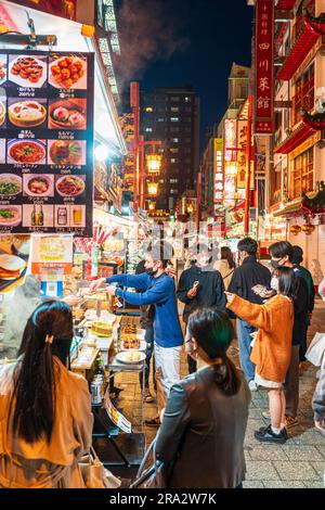 People waiting around a Chinese street stall selling steamed dumplings and buns and other Chinese food in the evening in Nankinmachi, Chinatown, Kobe. Stock Photo