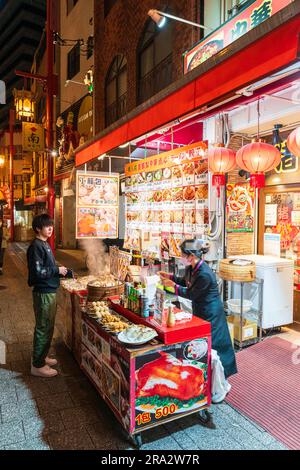 Woman serving young man at a Chinese food stall with steam rising from bamboo container of steamed buns. Night-time in Chinatown, Nankinmachi, Kobe. Stock Photo