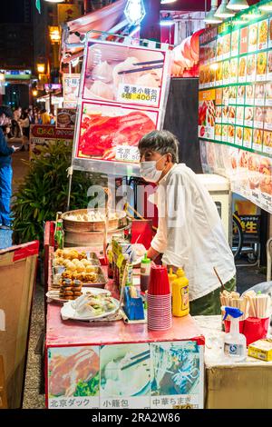 Side view of street vendor at his Chinese food stall sealing steamed dumplings and other foods, at night in Kobe's Chinatown, Nankinmachi. Stock Photo