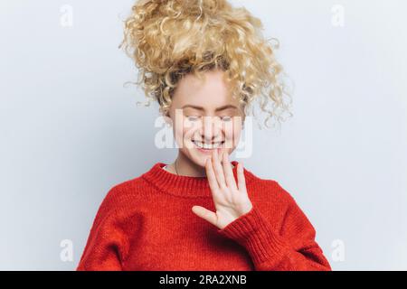 Happy young woman in red sweater, eyes closed, smiling, gesturing joyfully. Studio shot, white background Stock Photo