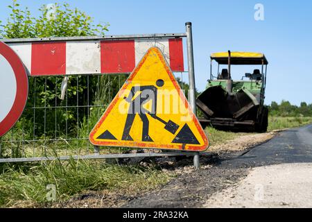 Road work ahead warning sign at construction site. Paver finisher equipment in bg. Crawler paving machine for laying asphalt. Roadworks traffic sign. Stock Photo
