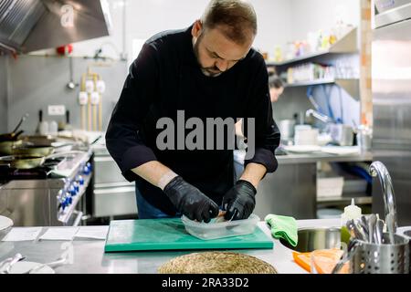 Bearded male professional chef in black gloves arraigning food into transparent container while working in professional kitchen Stock Photo
