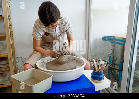 young ceramist on the lathe or potter's wheel working and shaping clay inside a pottery studio, creating creative shapes with tools. natural light Stock Photo